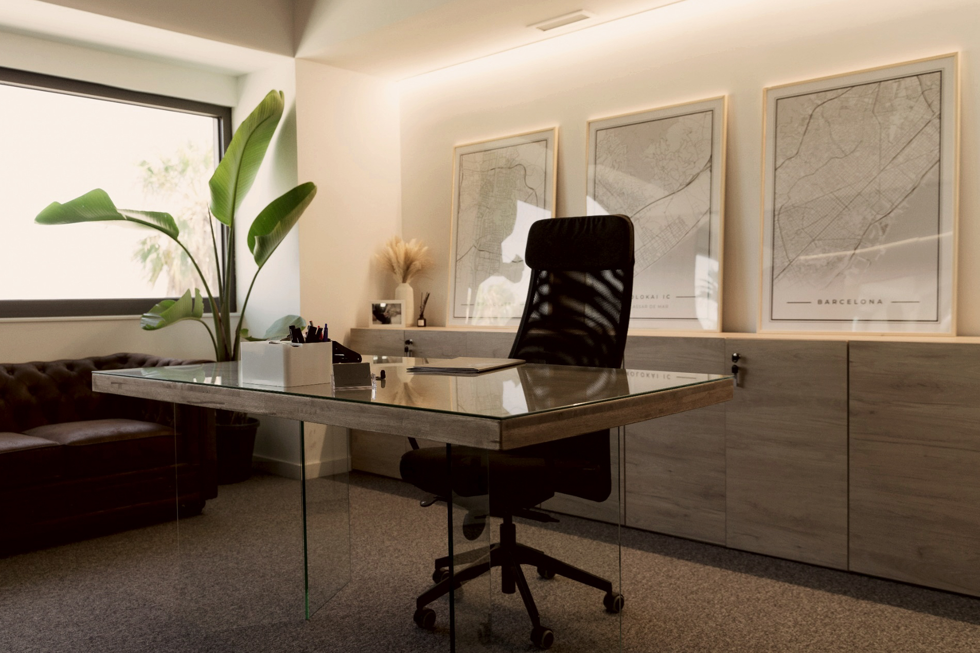 Despacho de Molokai con mesa de cristal, sillón de trabajo y planos de Barcelona en la pared